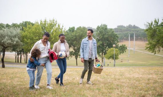 A joyful family walking through a park on a sunny day, carrying a picnic basket and a ball.