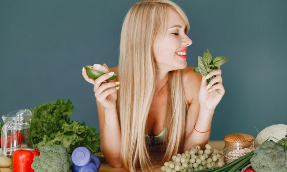 A smiling woman holding avocado and basil, surrounded by fresh vegetables and fruits for healthy eating.
